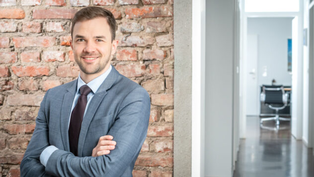 Mann im Anzug mit verschränkten Armen vor einer Backsteinwand in einem Büroflur