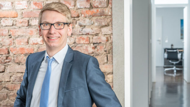 Mann in Anzug mit hellblauem Hemd und blauer Krawatte vor einer Ziegelsteinwand stehend