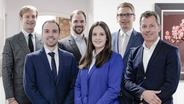 Sechs Personen in formeller Kleidung stehen in einem Raum mit weißen Wänden und einem Fenster im Hintergrund