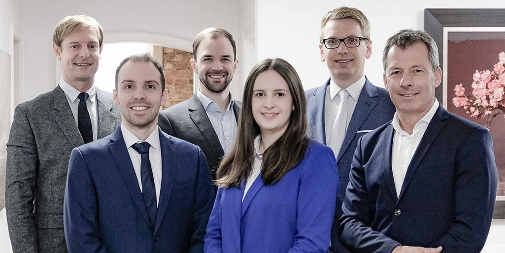 Sechs Personen in Businesskleidung stehen in einem Büro mit weißen Wänden und einem Bild mit rosa Blumen an der Wand