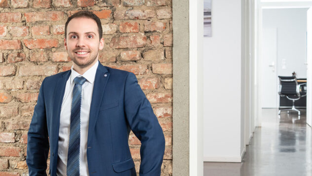 Mann in blauem Anzug mit weißem Hemd und gemusterter Krawatte lehnt an einer Ziegelsteinwand in einem modernen Büroflur