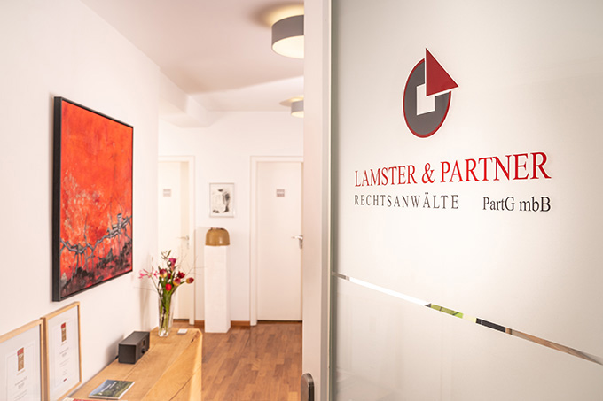 Blick in einen Flur mit Holzboden, links ein Sideboard mit Blumenstrauß und mehreren gerahmten Bildern, rechts eine Glastür mit rotem Logo und Schriftzug 'LAMSTER & PARTNER RECHTSANWÄLTE PartG mbB'