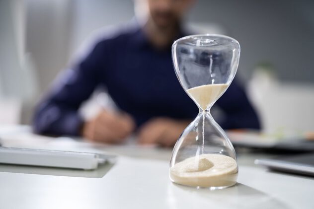 Sanduhr mit laufendem Sand auf einem Schreibtisch vor einer unscharfen Person, die schreibt