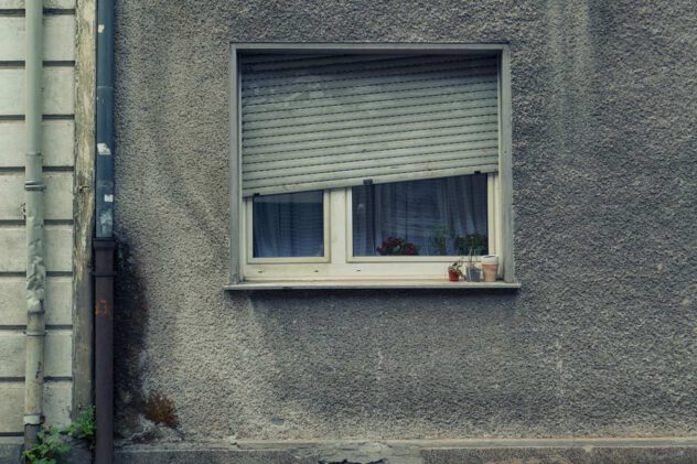 Fenster mit halb heruntergelassener Rolllade in einer rauen Putzfassade mit zwei kleinen Blumentöpfen auf der Fensterbank